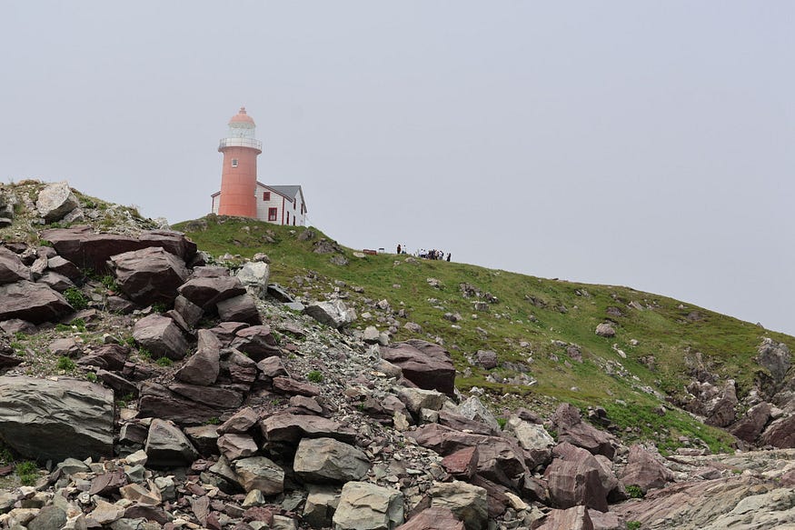 A lighthouse on a large hill is half-covered in fog. Several people, barely discernible, gather in a group around one another near the base of the lighthouse.