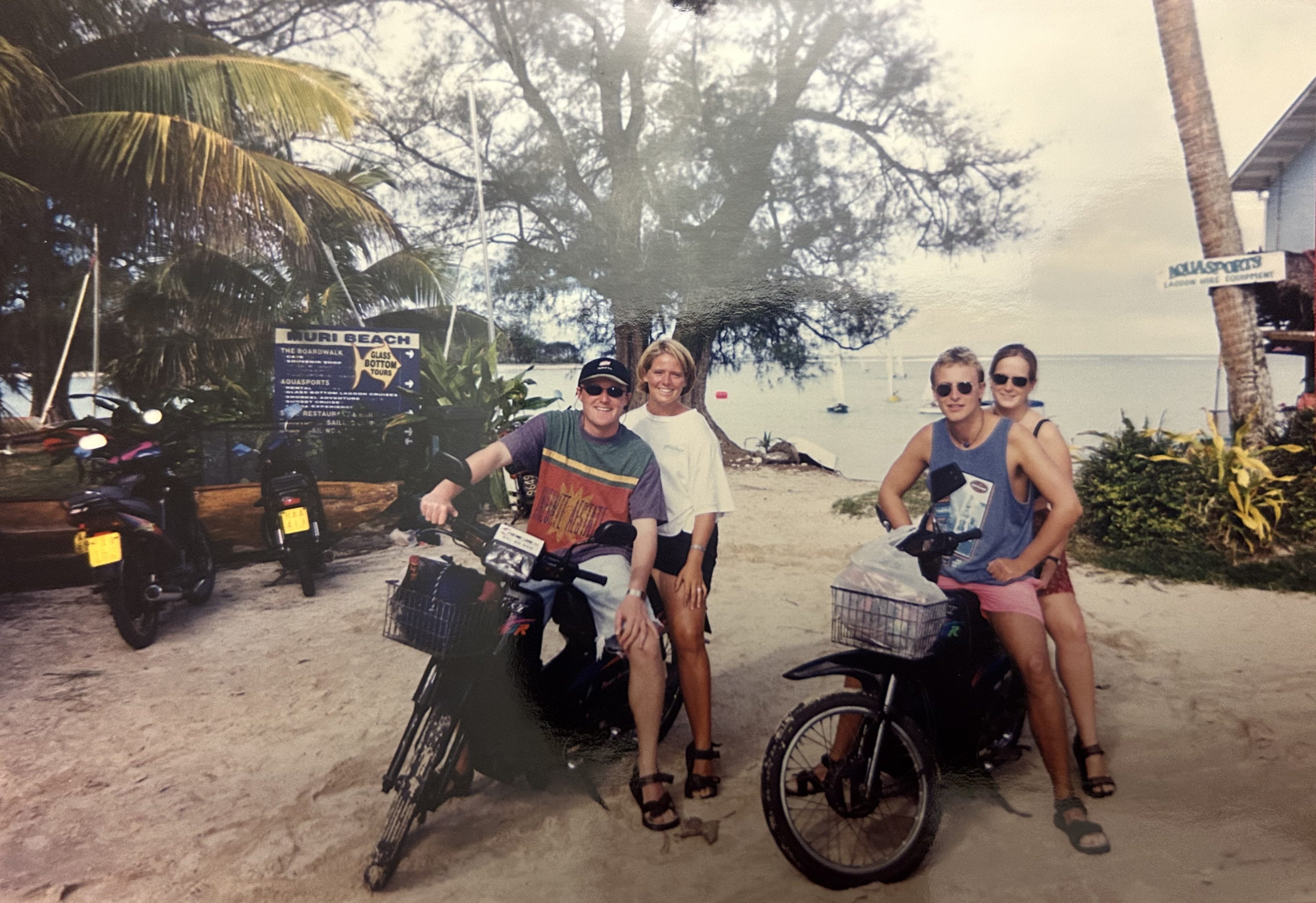 Two motorcycles with a man and woman on each. They are parked in the sand in front of the beach in the Cook Islands.