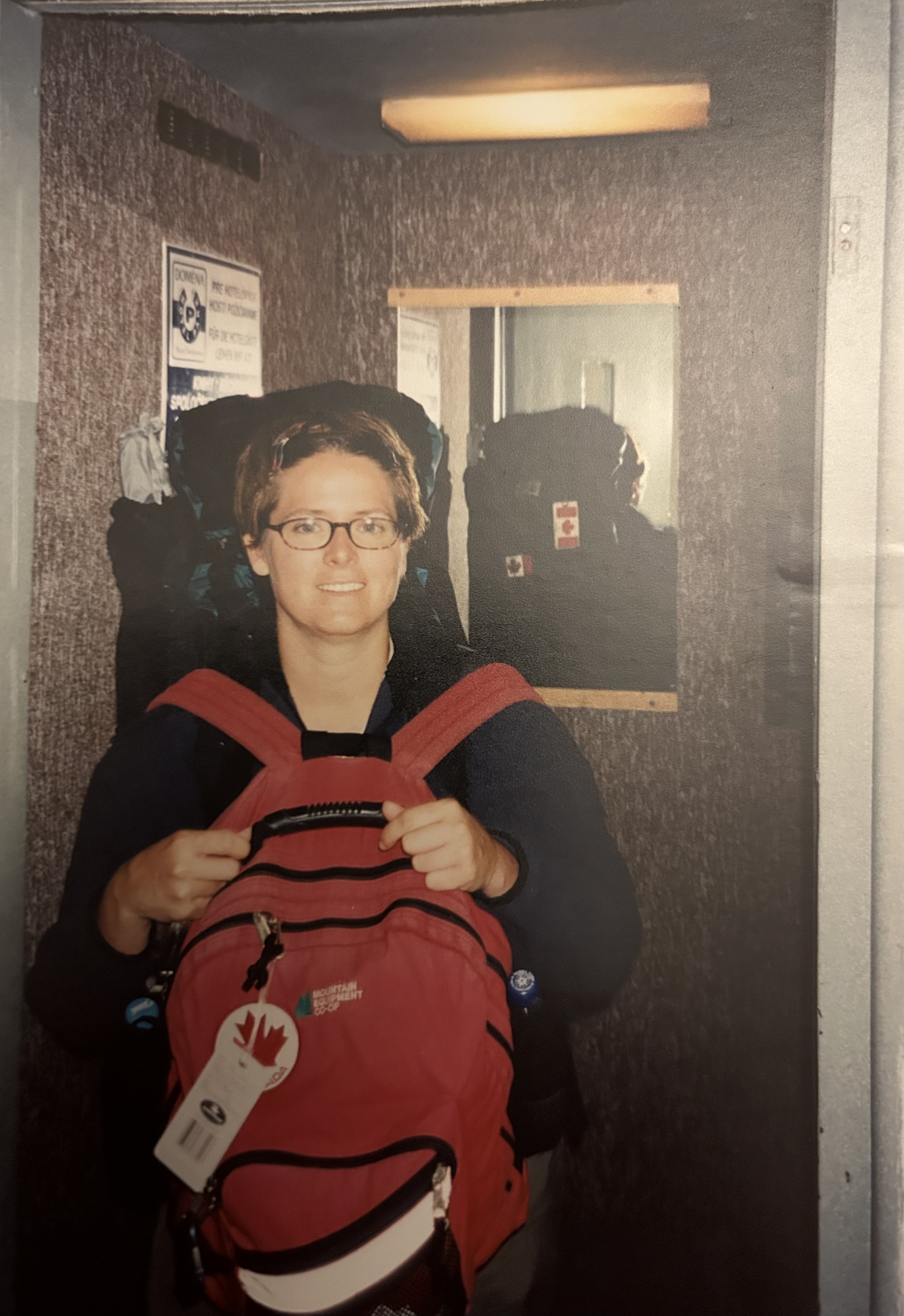 The author, a woman, wearing a large backpack standing in front of an elevator.  She has a red backpack strapped on the front as well.