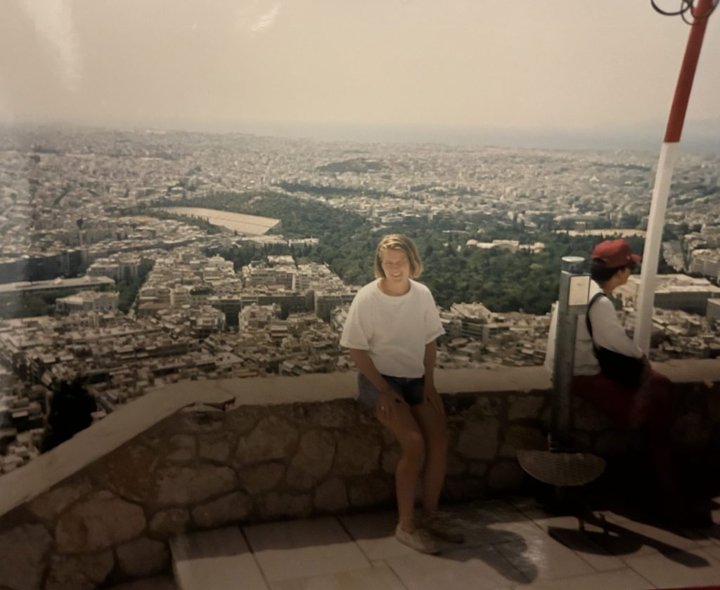 The author as a teenager with short blond hair wearing a white shirt and jeans shorts. She is sitting on a stone wall overlooking the city of Athens, Greece, in 1992.