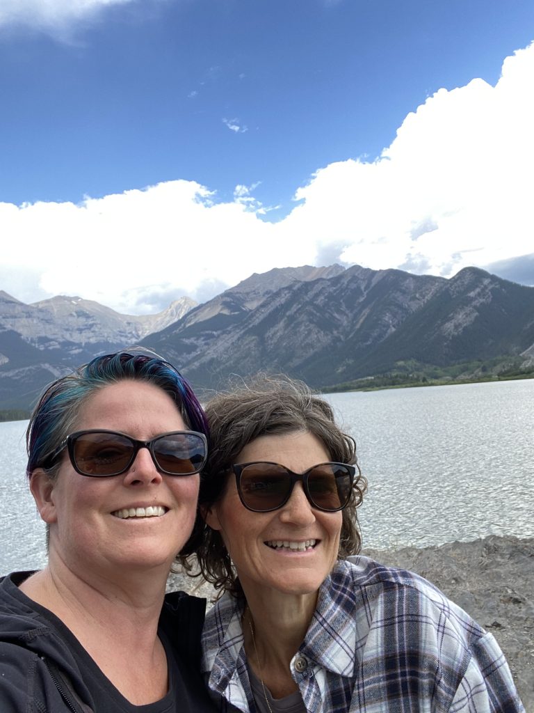 Two women wearing sunglasses stand in front of a lake and mountain.