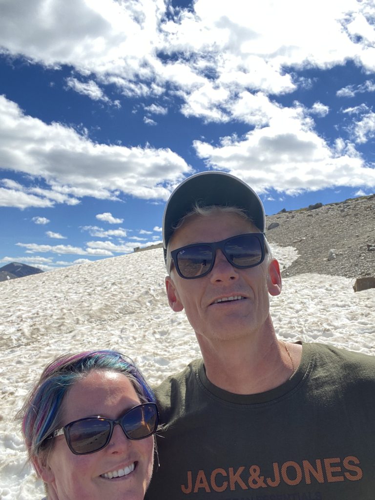 A couple standing on the top of a mountain with snow behind them.